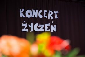 Fotografia przedstawiająca duży, biały napis '' Koncert Życzeń", który umieszczony jest na czarnym tle. Na pierwszym planie rozmazane kolorowe kwiaty.