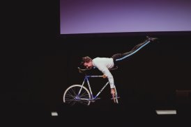 Wnętrze Teatru. Na scenie występuję cyrkowiec, który prezentuję różne triki na rowerze. Mężczyzna jest przebrany w białą koszulę i czarne spodnie.