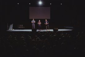 Wnętrze Teatru. Na scenie trzech mężczyzn prezentujących swój pokaz. Publiczność wpatruje się w nich z zaciekawieniem. Mężczyźni są ubrani w kolorowe stroje.