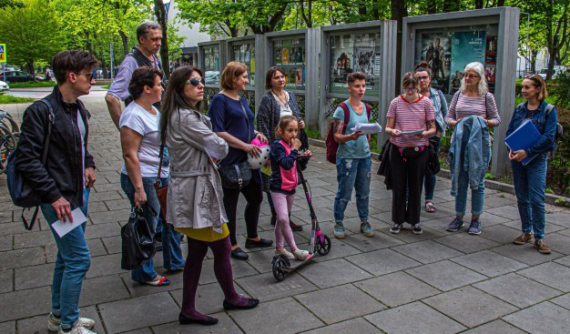 Zdjęcie przedstawiające grupę kilkunastu osób w różnym wieku, stojącą gdzieś na chodniku w otoczeniu zieleni