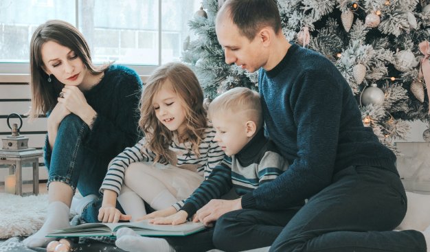 Rodzina, mama tata i dwójka dzieci - chłopiec i dziewczynka, siedzą na podłodze blisko ubranej choinki bożonarodzeniowej. Wspólnie przeglądają książkę.