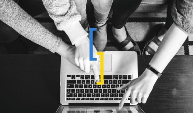 Widok z góry na ręce kilku osób, które siedzą przy laptopie i wskazują coś rękoma na ekranie komputera. Centralnie umieszczone dwa zamknięte nawiasy w kolorach niebieskim i żółtym.