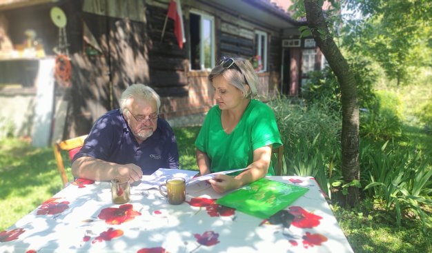 Zdjęcie przedstawiające dwie osoby siedziące przy stole na podwórku. W tle widoczne są drzewa i krzaki oraz drewniany dom z flagą biało-czerwoną. Przy stole siedzi starszy mężczyzna i kobieta w zielonej bluzce, pochylają się nad papierami leżącymi na stole.