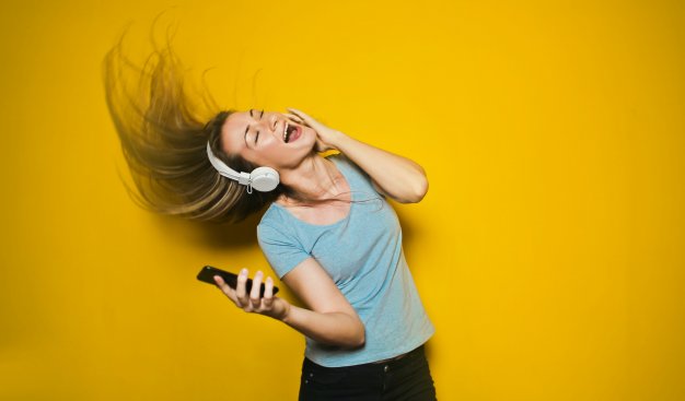 Młoda kobieta w słuchawkach i z telefonem w ręku, tańczy na tle żółtej ściany.