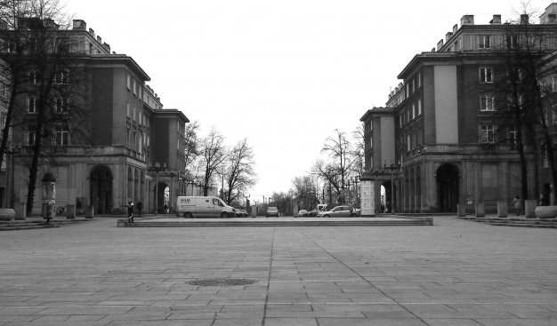 Czarno-białe zdjęcie Alei Róż w kierunku Placu Centralnego. Pochmurny dzień, brak ludzi. W tle drzewa bez liści, przed nimi pojazdy stojące na parkingu w osi szerokiej alei. Po bokach budynki mieszkalne z charakterystycznymi arkadami. W dole zdjęcia płyty