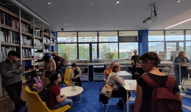 Jasne, nowoczesne wnętrze biblioteki, utrzymane w tonach popieli i błękitu. Na ścianie duże okna ciągnące się przez całe pomieszczenie, na bocznej - półki z książkami. W pomieszczeniu liczna grupa osób w różnym wieku i różnej płci. To gracze drugiej rozgr
