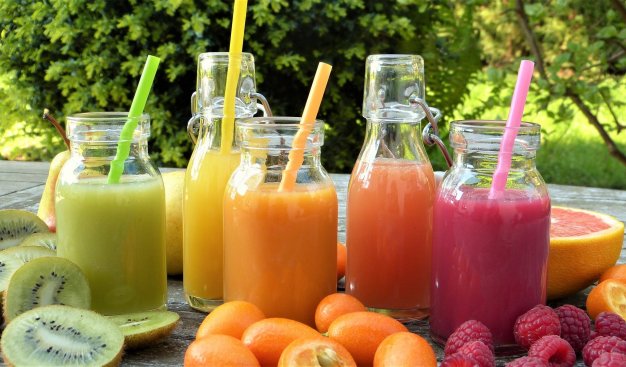 Szklane butelki z kolorowymi sokami owocowymi, wokół butelek leżą owoce: kiwi, pomarańcze, maliny.