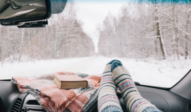 Zimowy dzień. Widok z wnętrza samochodu na drogę przez las. Ziemi i drzewa pokryte śniegiem. Na pierwszym planie nogi w skarpetach na podszybiu auta, obok leży koc z książką.