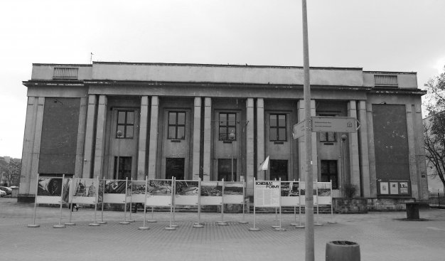 Czarno-białe zdjęcie przedstawiające byłe Kino Światowid - obecnie Muzeum PRL'u. Przed nim rząd kilkunastu tablic przedstawiających archiwalne zdjęcia z Kombinatu.