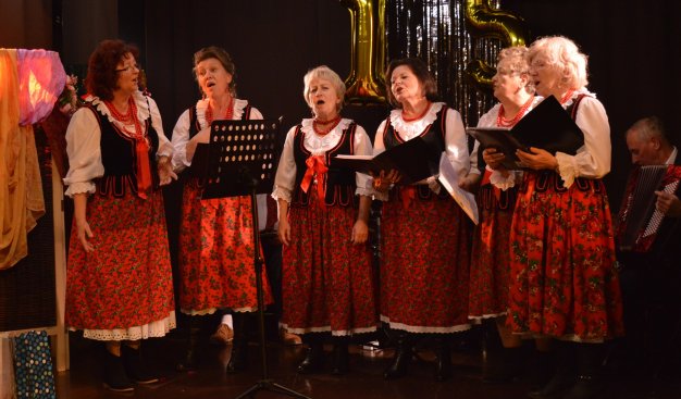 Zespół woklany kobiet seniorek ubranych w ludowe stroje krakowskie. Śpiewają, trzymając w rękach teczki z nutami.
