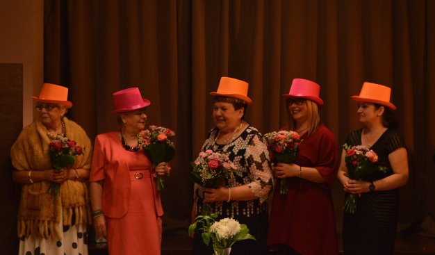 Pięć kobiet w kolorowych kapeluszach stoi w rzędzie, w rękach trzymają niewielkie bukiety kwiatów.