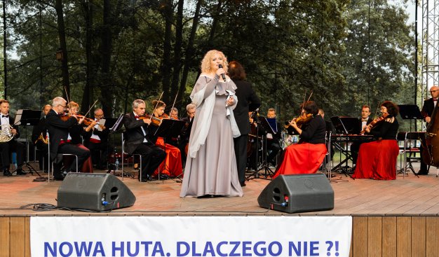 Zdjęcie przedstawiające kobietę - Lidię Jazgar stojącą na scenie, na dole zdjecia znajduje się baner z napisem Nowa Huta. Dlaczego nie?!