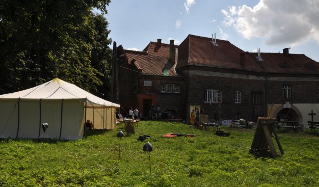widok na Klub Aneks w słoneczny dzień. Na pierwszym planie zielona trawa i biały namiot, oraz porozkładane sprzęty z epoki średniowiecznej.