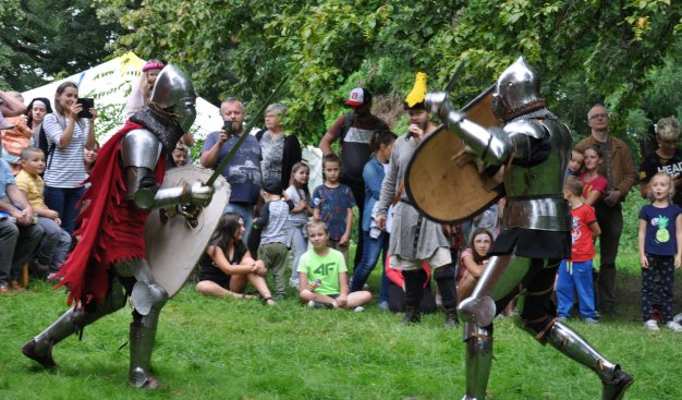 Widok na walkę na miecze i tarcze dwóch osób w średniowiecznych zbrojach. Wokół nich liczna widownia. Cała scena na zielonej trawie