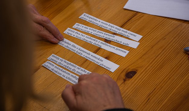 Zdjęcie pzredstawiające stół na którym znajdują się pocięte w paski kartki, na których znajdują się fragmenty tekstów wierszy. Na pierwszym planie widoczne są ręce uczestników warsztatów, które układają pocięte fragmenty w odpowiedniej kolejności.
