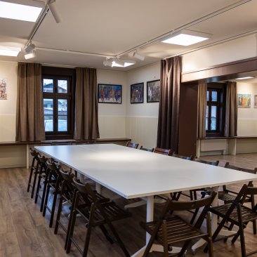 Zdjęcie przedstawiające jasne wnętrze Klubu Krzesławice. Na środku pomieszczenia duzy stół z klkudziesięcioma krzesłami wokół.